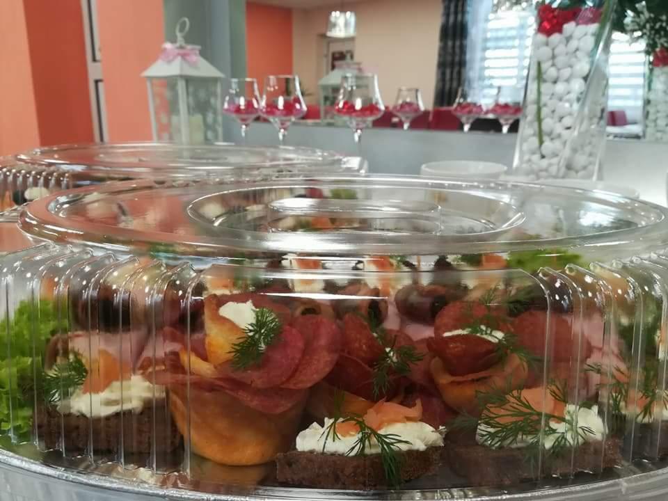 Przekąski bankietowe w przezroczystych pojemnikach, widoczne kanapki z ciemnego pieczywa z kremowym serem, wędlinami i koperkiem, oraz babeczki z salami, w tle dekoracje stołu z lampionem...