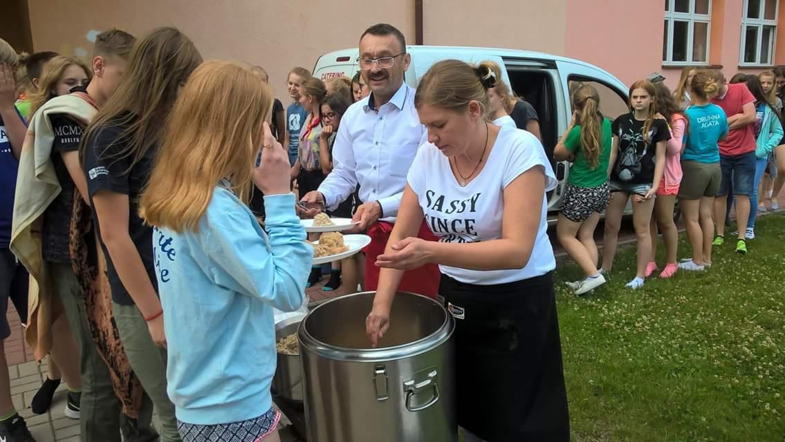 Dystrybucja posiłku z dużego metalowego gara przez kobietę w czarnym fartuchu, z mężczyzną w białej koszuli i okularach trzymającym talerze, oraz kolejką młodzieży w tle, prawdopodobnie podczas...