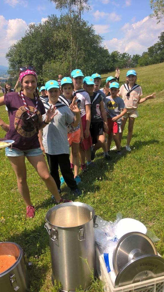 Grupa dzieci w jednakowych czapkach ustawiona w rzędzie na zielonej łące, z przodu duże metalowe garnki z jedzeniem i pokrywki, prawdopodobnie na pikniku skautowym.