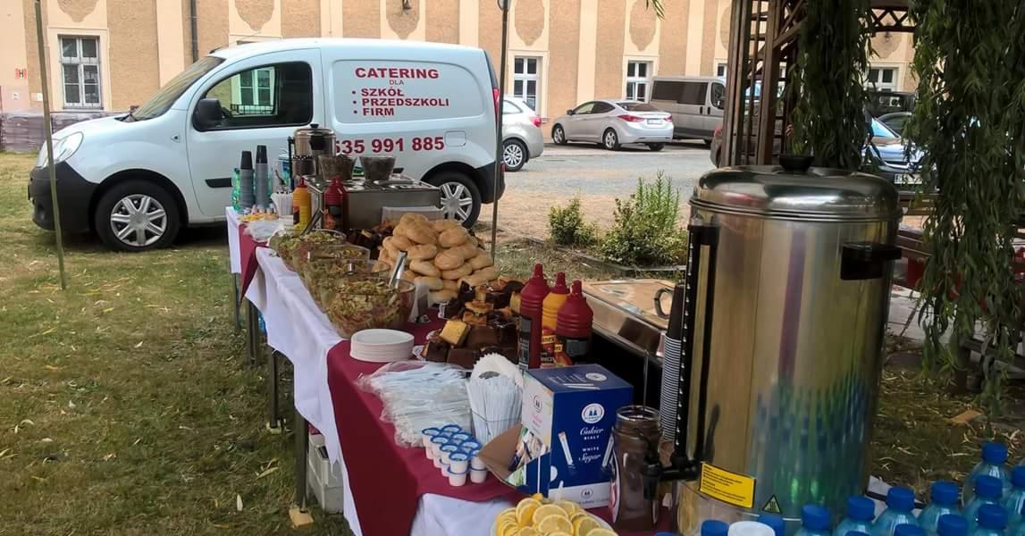 Stół z różnorodnym jedzeniem i napojami na zewnątrz, obok zaparkowany biały samochód dostawczy z napisem 'Catering dla szkół, przedszkoli, firm' i numerem telefonu, budynek w tle.