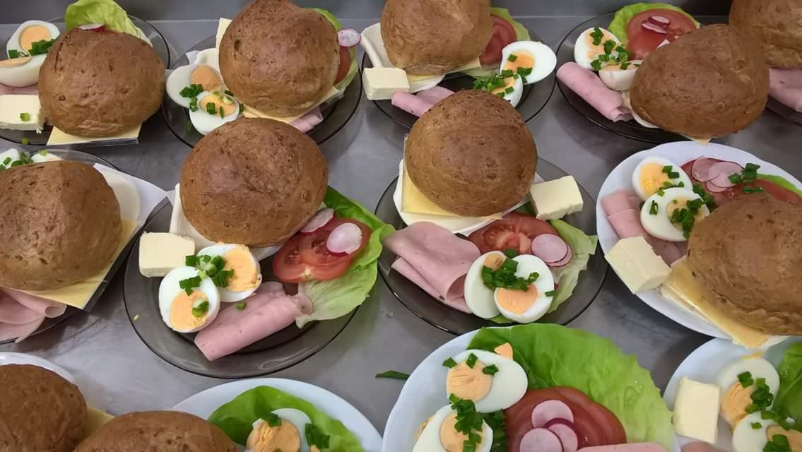 Porcje śniadaniowe na talerzach: bułka z serem, szynką i warzywami, jajko na twardo posypane szczypiorkiem, plaster masła, pomidor i rzodkiewka.