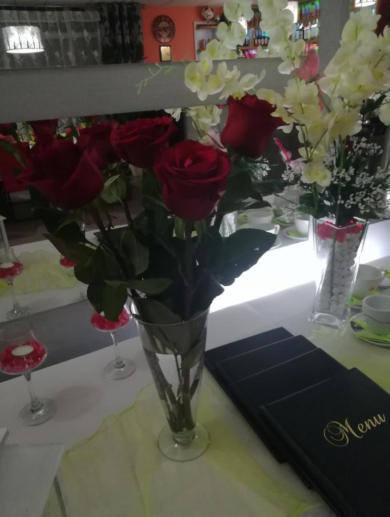 Dekoracja stołu z bukietem czerwonych róż w szklanym wazonie, obok menu w czarnej oprawie ze złotym napisem, oraz kieliszkami z dekoracyjnymi kamyczkami na tle białych kwiatów w wazonie.