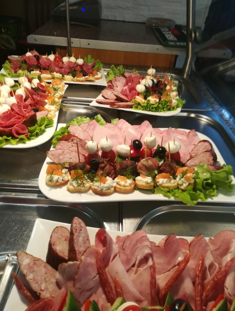 Szeroki kadr ukazujący kilka białych pater z różnorodnymi przekąskami: wędliny, sery mozzarella na wykałaczkach z pomidorkami, mini kanapeczki z pastami, wszystko udekorowane zieloną sałatą.