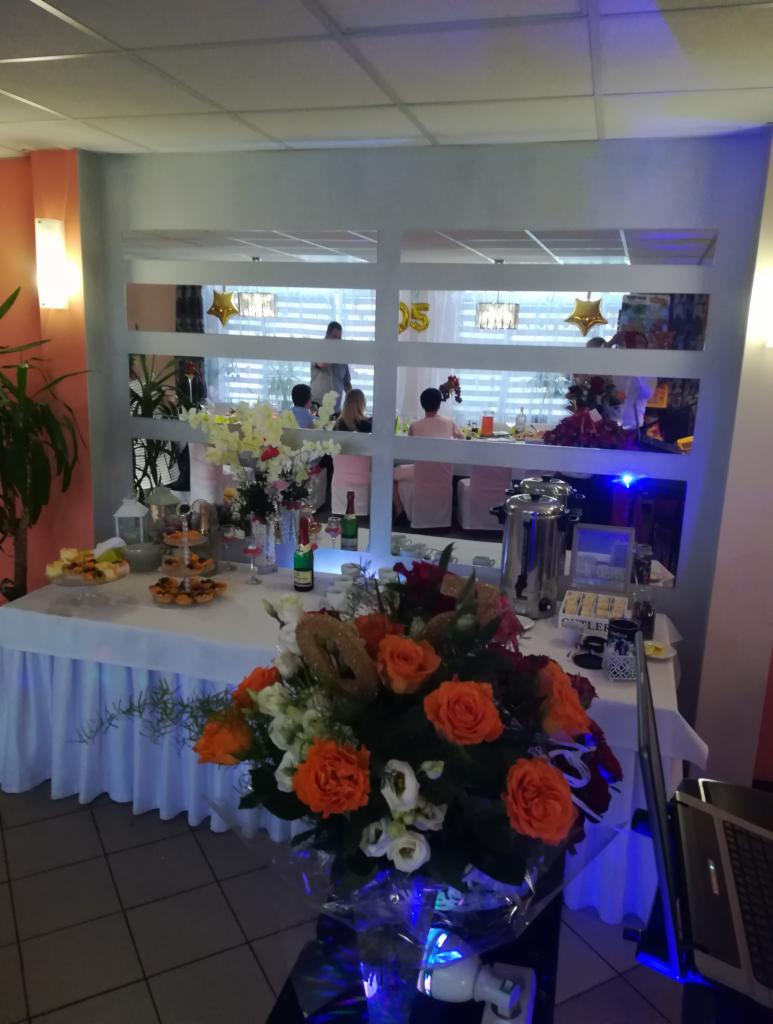 Stół z przekąskami, napojami i dekoracjami kwiatowymi przed salą z gośćmi i balonami w tle, ustawiony podczas imprezy okolicznościowej.