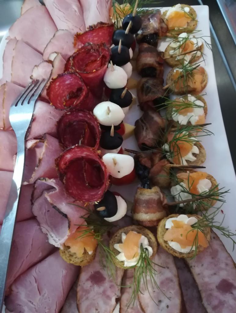 Dekoracyjna taca z przekąskami: wędliny, koreczki z mozzarelli i oliwek, śliwki w boczku oraz mini kanapeczki z łososiem i kremowym serkiem, udekorowane gałązkami rozmarynu i widelcem.