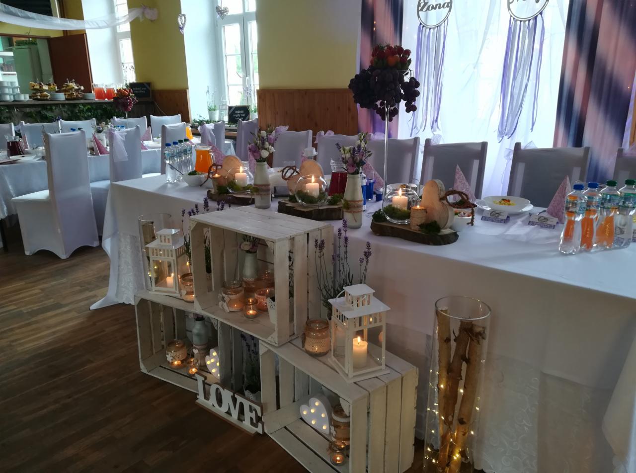 Uroczysta dekoracja stołu weselnego z białym obrusem, drewnianymi skrzynkami z lampionami i świecami, kwiatami w wazonach, szklanymi kulami ze świecami, oraz dekoracją 'Zona' w tle.