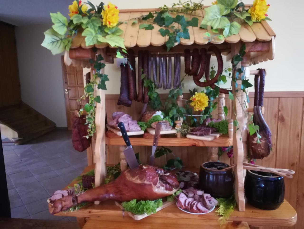 Dekoracyjna drewniana konstrukcja prezentująca wędliny, kiełbasy i szynki, ozdobiona żółtymi kwiatami i zielonymi liśćmi, z nożem wbitym w szynkę na pierwszym planie.