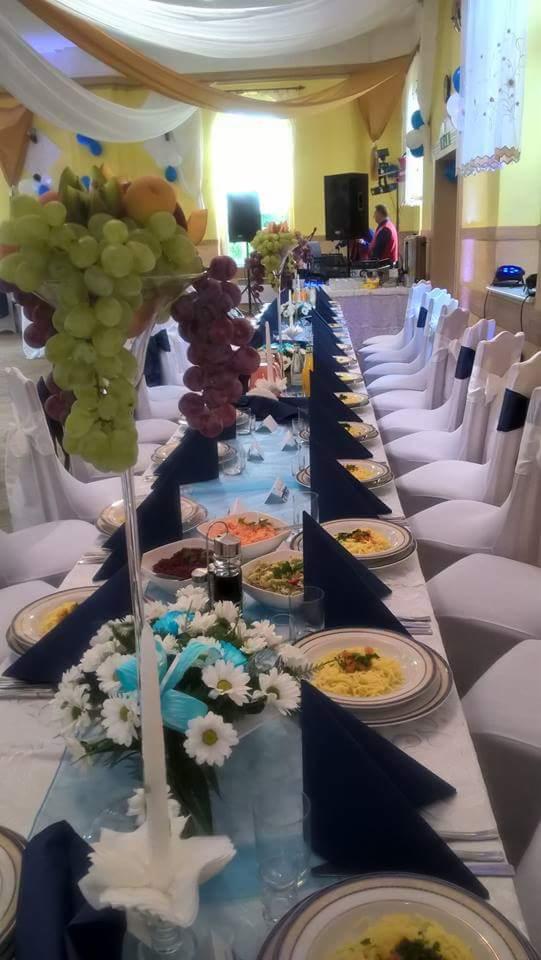 Długi stół bankietowy z białymi krzesłami udekorowany owocami, kwiatami, ciemnoniebieskimi serwetkami i zastawą stołową z daniami, w tle dekoracje sali z białych i złotych tkanin.