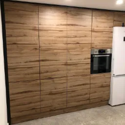 Kuchenna zabudowa z drewnopodobnymi frontami, czarne obramowanie, piekarnik w zabudowie, biała lodówka obok.