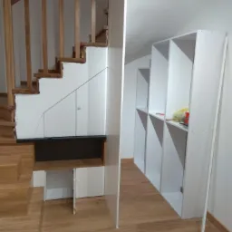 Biała zabudowa pod schodami z szafkami i półkami, drewniane schody z balustradą, jasna podłoga, częściowo zagospodarowana przestrzeń.