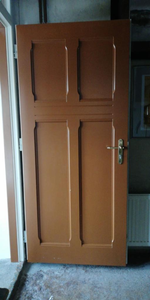 Brązowe drzwi wewnętrzne z czterema panelami, widoczne zawiasy i klamka, surowe wykończenie podłogi.