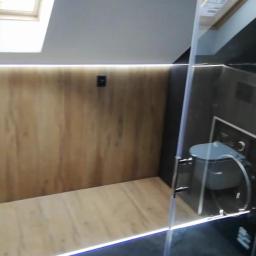 Firma ogólnobudowlana Marcin Zabagło - Nowoczesna łazienka na poddaszu z drewnopodobnymi płytkami na ścianach i podłodze, oświetleniem LED przy podłodze i suficie, oraz kabiną prysznicową z czarnymi elementami i widoczną toaletą.