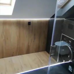 Nowoczesna łazienka na poddaszu z drewnopodobnymi płytkami na ścianach i podłodze, oświetleniem LED przy podłodze i suficie, oraz kabiną prysznicową z czarnymi elementami i widoczną toaletą.