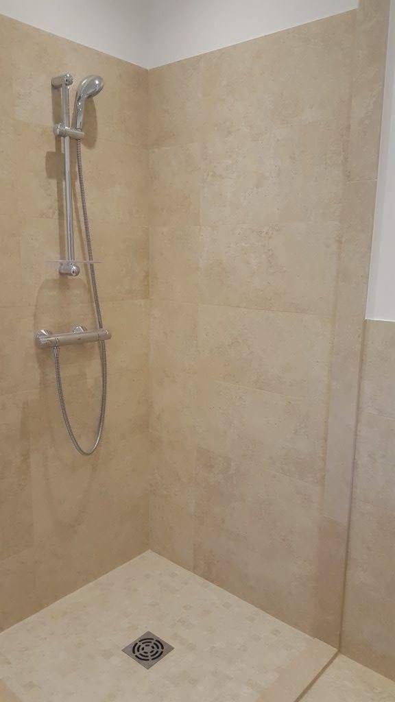 Wykończony narożnik kabiny prysznicowej wyłożony beżowymi płytkami imitującymi kamień, z chromowaną baterią i słuchawką prysznicową oraz odpływem liniowym w podłodze.