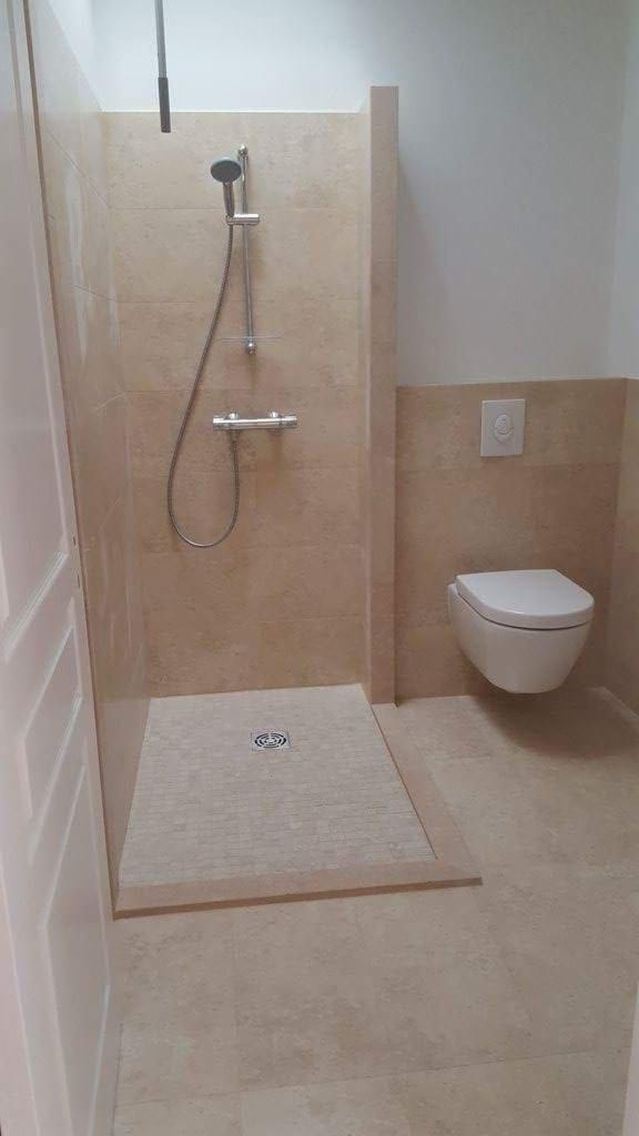 Beżowa łazienka z kabiną prysznicową walk-in wyłożoną płytkami imitującymi kamień, widoczny odpływ liniowy i wisząca toaleta.