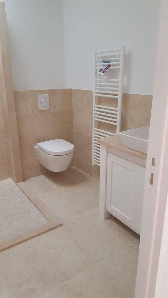 Beżowa łazienka z podwieszaną toaletą, grzejnikiem drabinkowym i białą szafką pod umywalkę z blatem.