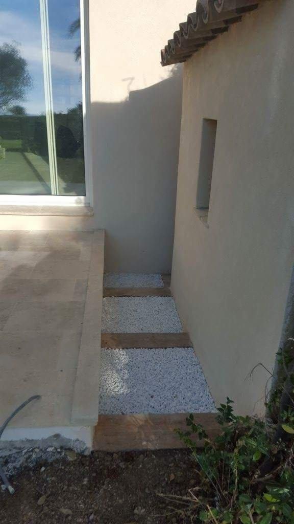 Ułożone kamienne ścieżki z drewnianymi belkami oddzielającymi fragmenty białego żwiru, prowadzące wzdłuż ściany budynku z oknem i małym otworem okiennym.