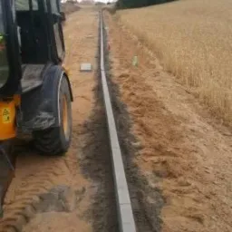 Żółta koparko-ładowarka częściowo widoczna po lewej stronie zdjęcia, obok ułożone krawężniki oddzielające drogę od pola w słoneczny dzień.