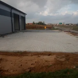 Nowa nawierzchnia z szarej kostki brukowej ułożona przed budynkiem magazynowym z widocznym przygotowanym podłożem z ziemi i oddalonym krajobrazem wiejskim.