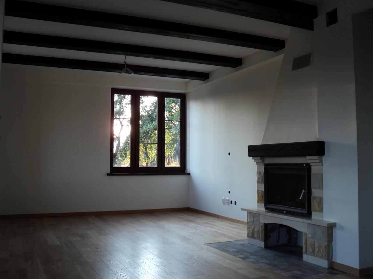 Jasny salon z parkietem, oknem z widokiem na zieleń i kominkiem z kamienia. Sufit z ciemnymi belkami. Nowoczesne wykończenie wnętrza.