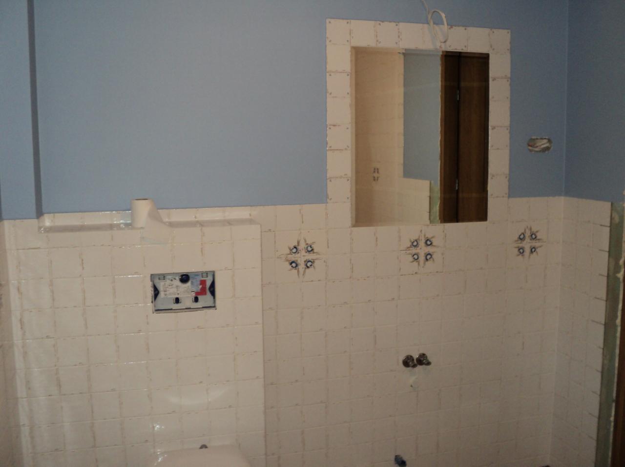 Błękitna ściana nad białymi, kwadratowymi płytkami w łazience. Widoczne przygotowania do montażu armatury i lustra. Charakterystyczny wzór na fugach płytek.