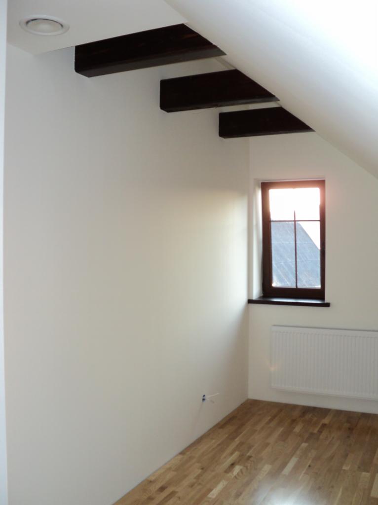 Jasne wnętrze poddasza z drewnianą podłogą, oknem w ciemnej ramie i białymi ścianami. Widoczne drewniane belki stropowe i grzejnik.
