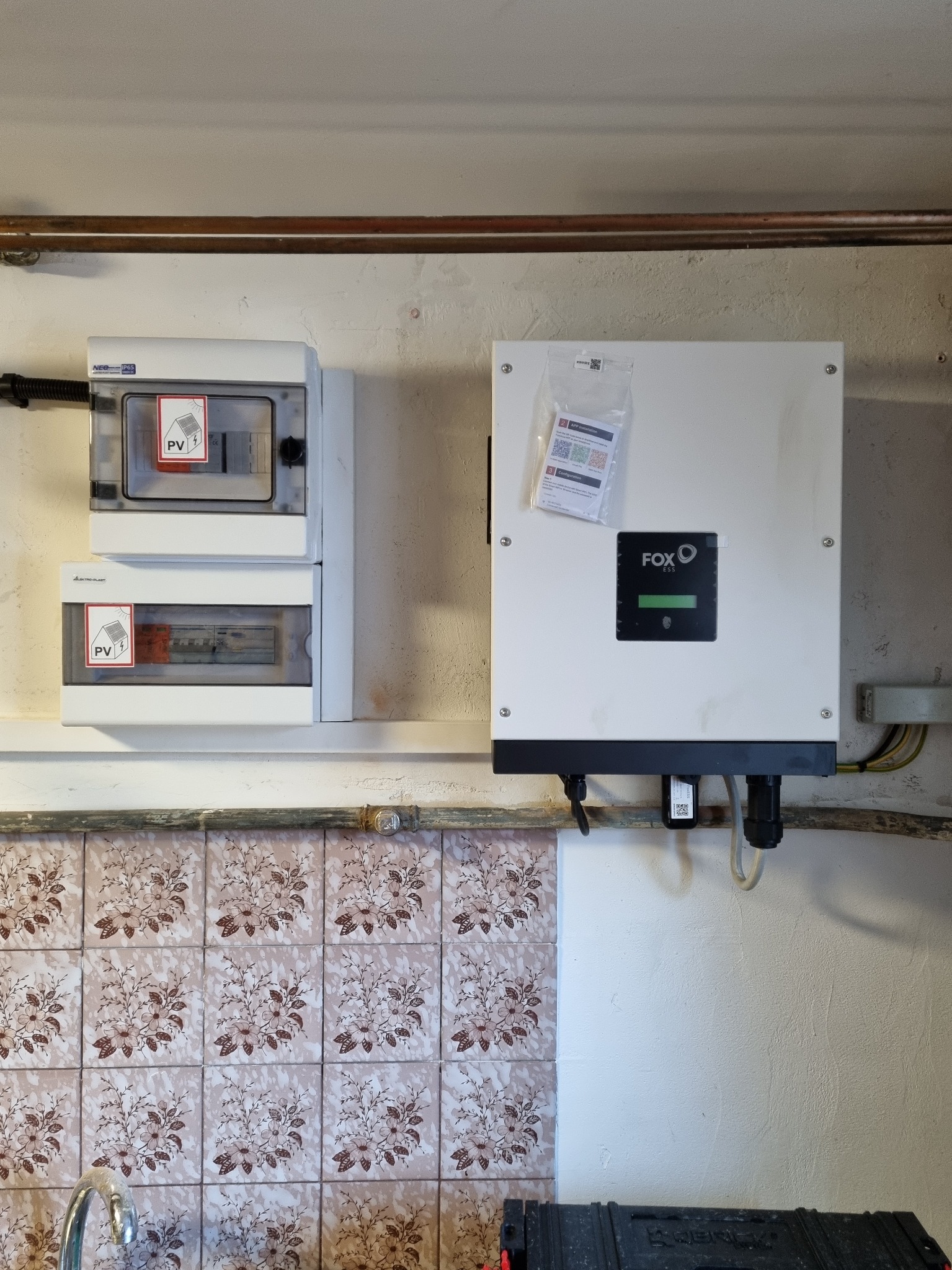 Biała skrzynka inwertera z logo FOX ESS zamontowana na ścianie nad staromodnymi, wzorzystymi kafelkami, obok dwie mniejsze skrzynki z naklejkami PV.