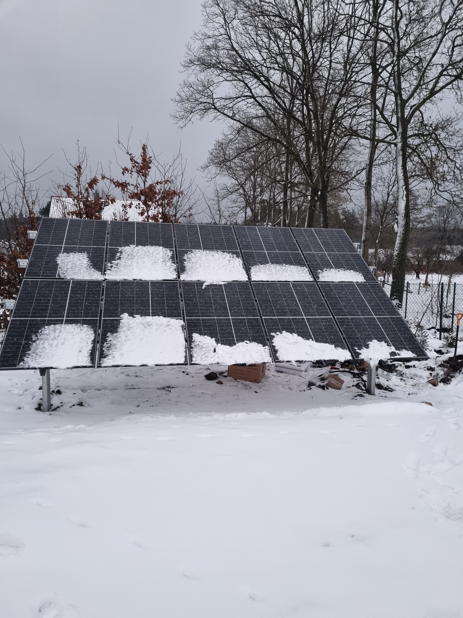 Panele słoneczne częściowo pokryte śniegiem, zamontowane na metalowych wspornikach, na tle zaśnieżonego terenu i drzew bez liści w pochmurny dzień.