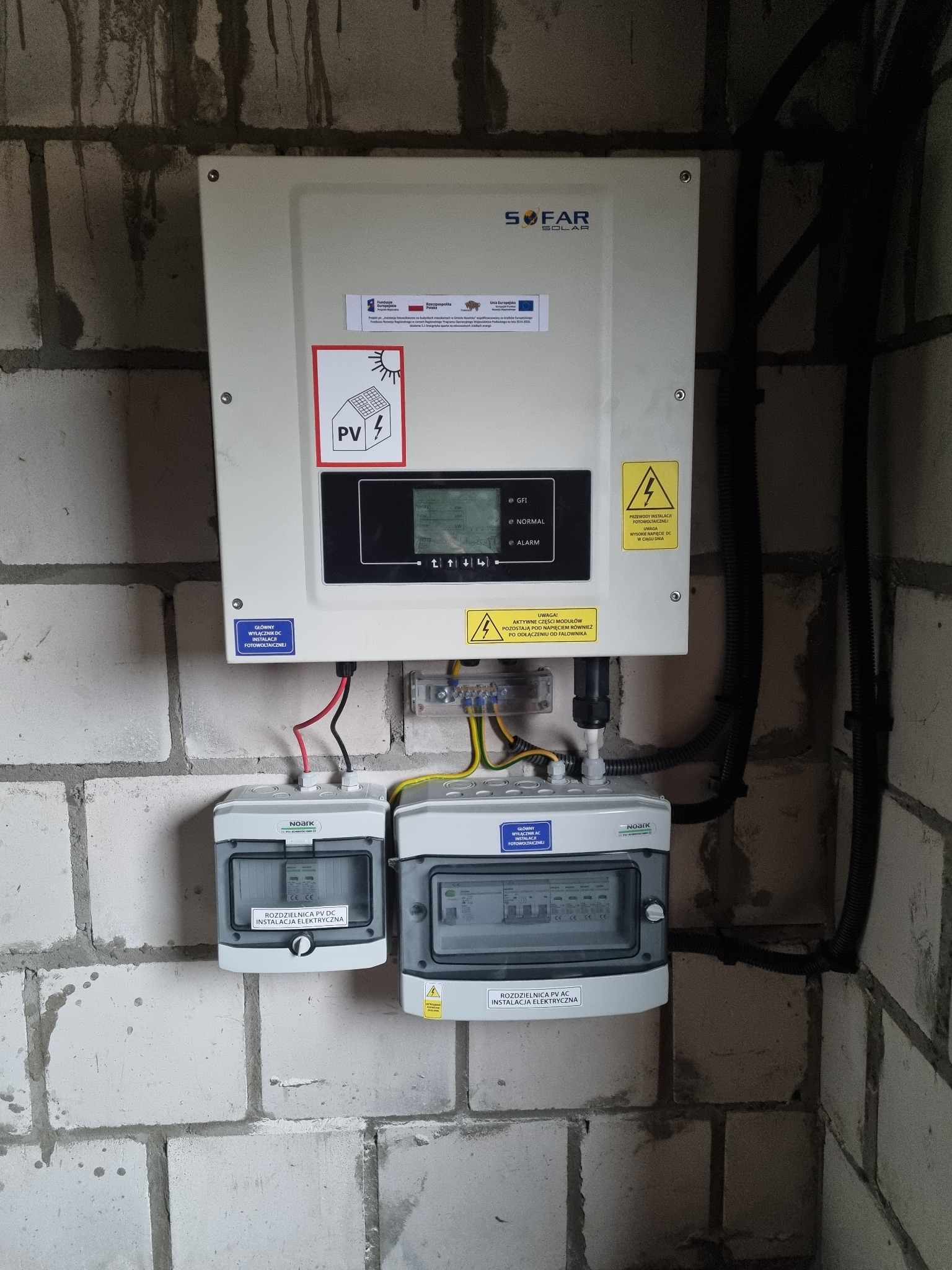Zamontowany na surowej ścianie falownik SOFAR SOLAR z rozdzielnicami DC i AC, okablowanie w peszlach. Widoczne naklejki ostrzegawcze i informacyjne dotyczące instalacji fotowoltaicznej.