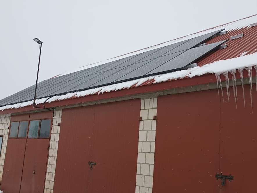 Panele fotowoltaiczne pokryte śniegiem na czerwonym dachu garażu z wiszącymi soplami lodu, oświetlone lampą uliczną.