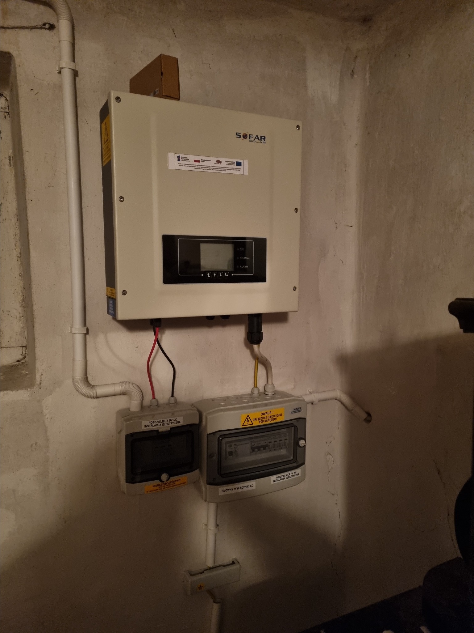 Zamontowany inwerter Sofar Solar z podłączonymi rozdzielnicami DC i AC w pomieszczeniu o surowych, betonowych ścianach. Widoczne przewody i rury instalacyjne.