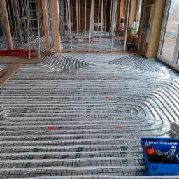 Białe rurki instalacji grzewczej ułożone spiralnie na podłodze w surowym stanie, zabezpieczone folią, widoczny niebieski koszyk z narzędziami i fragment konstrukcji ścian.