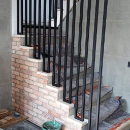 Reflex - Nowoczesne schody z czarną, metalową balustradą o prostych, pionowych szczeblach, zabezpieczone na czas budowy, z fragmentem ściany wykończonej czerwoną cegłą i szarą płytą betonową w tle.