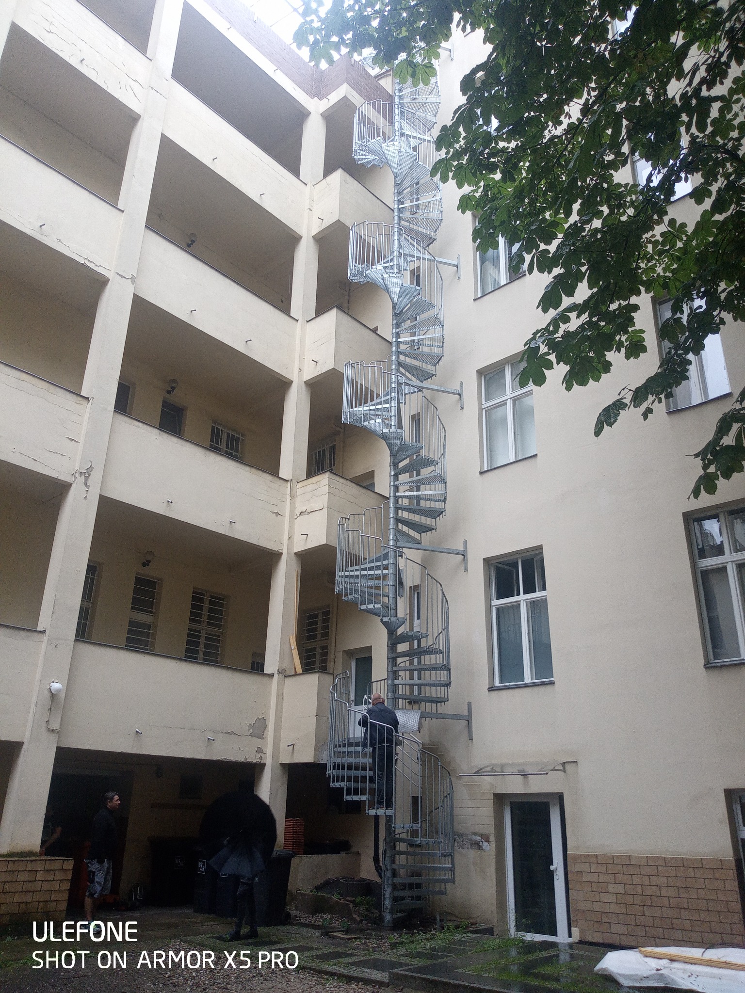 Zewnętrzna, metalowa klatka schodowa spiralna przymocowana do ściany budynku mieszkalnego z balkonami, osoba wchodzi po schodach.