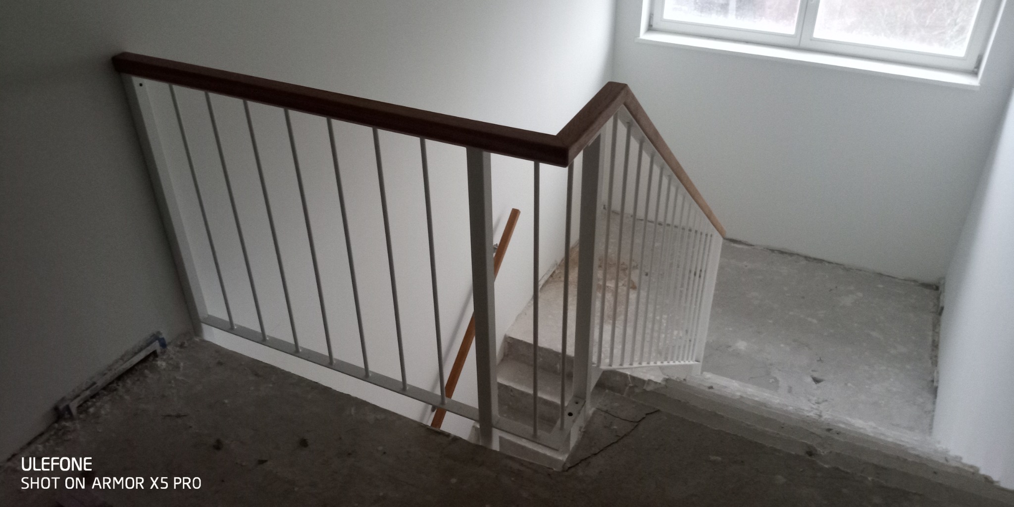 Metalowa balustrada schodowa z drewnianym poręczą w surowym stanie, widok z góry na betonowe schody w trakcie remontu.