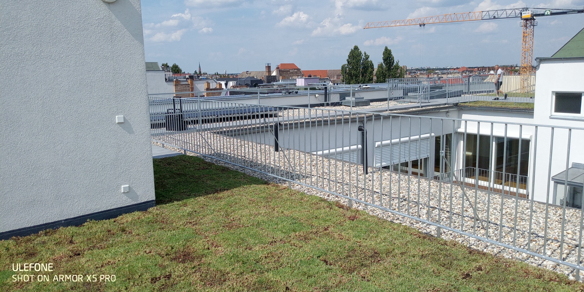 Metalowe balustrady zabezpieczające zielony dach budynku z widokiem na miasto i dźwig budowlany w tle.