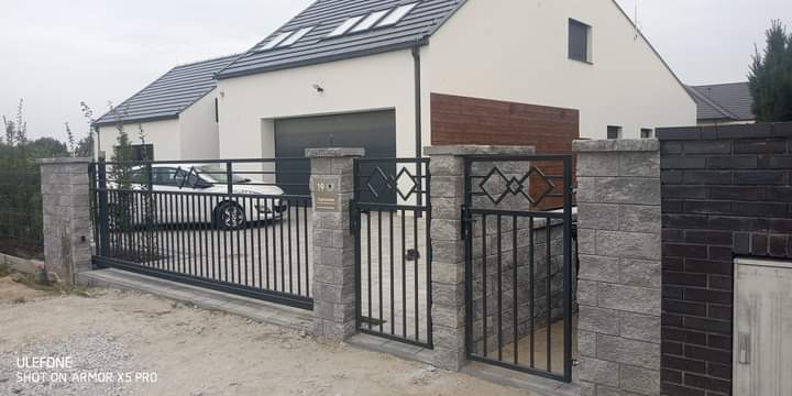 Nowoczesna posesja z przesuwną bramą wjazdową i furtką, czarne metalowe elementy z geometrycznymi zdobieniami, słupki z szarej kostki, w tle biały dom z garażem i fragment ciemnego ogrodzenia.