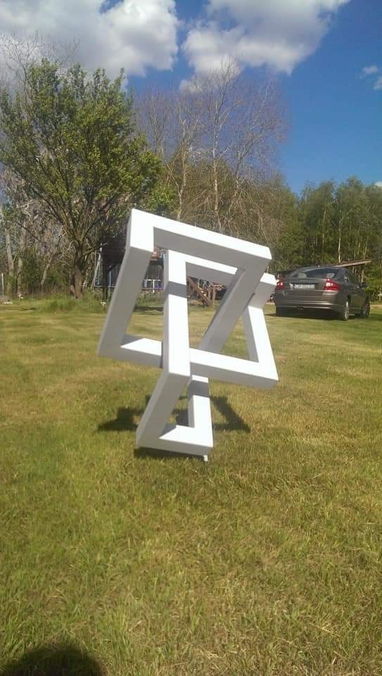 Biała, przestrzenna konstrukcja w formie niemożliwego trójkąta Penrose'a, ustawiona na trawniku przed domem jednorodzinnym, z samochodem osobowym w tle.