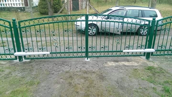 Zielona, dwuskrzydłowa brama z automatycznym napędem, zdobiona kutymi elementami, na tle szarego kombi.