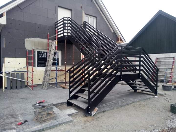 Zewnętrzne, metalowe schody z czarną balustradą prowadzące na balkon, widoczne podczas montażu przy ocieplonym budynku, z rozstawionymi rusztowaniami i narzędziami.