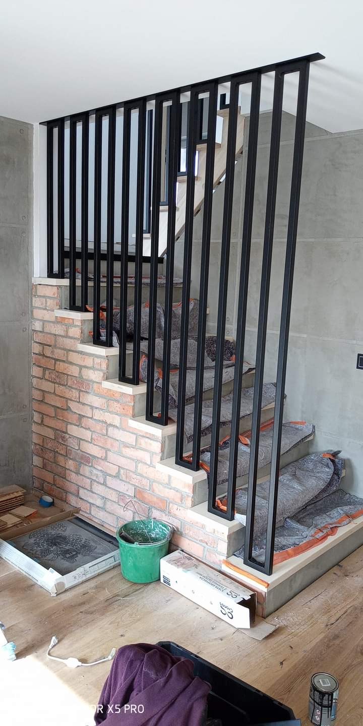 Nowoczesne schody z czarną, metalową balustradą o prostych, pionowych szczeblach, zabezpieczone na czas budowy, z fragmentem ściany wykończonej czerwoną cegłą i szarą płytą betonową w tle.