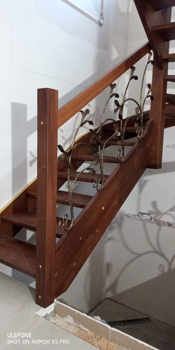 Drewniane schody z dekoracyjną, metalową balustradą w kształcie stylizowanych pnączy z liśćmi, widoczne niedokończone prace wykończeniowe przy podłodze.
