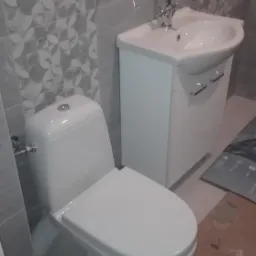 Biała toaleta i umywalka z szafką w łazience wykończonej szarymi płytkami z geometrycznym wzorem, widoczne wystające kable elektryczne.