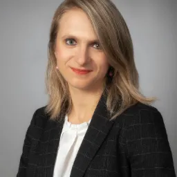 Portret uśmiechniętej kobiety w czarnej marynarce w kratę i białej bluzce na szarym tle, z blond włosami opadającymi na ramiona i różowymi kolczykami.