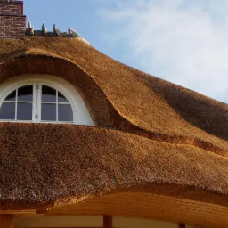 Strzecha z okrągłym oknem i ceglanym kominem na tle błękitnego nieba.