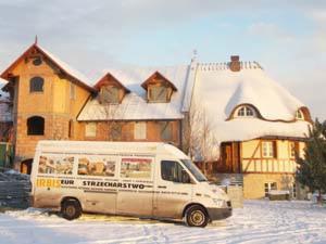 Zimowy krajobraz w Świerznie: zabytkowy budynek z unikalnym, śnieżnym dachem w stylu strzechy, częściowo oświetlony popołudniowym słońcem. Na pierwszym planie zaparkowany biały bus z reklamą firmy...