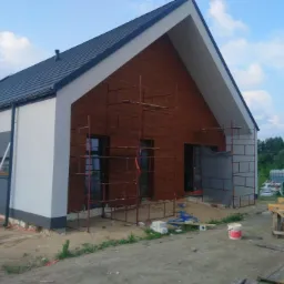 Nowoczesny dom jednorodzinny w trakcie wykańczania elewacji, widoczne rusztowania, fragmenty ocieplenia i pomalowane ściany w odcieniach szarości i brązu.