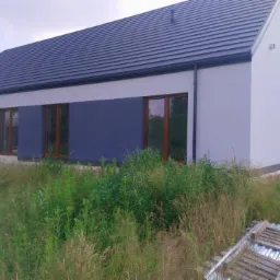 Nowoczesny dom murowany z dwukolorową elewacją (biało-granatową) i ciemną dachówką, widok z zarośniętego ogrodu, z paletami na pierwszym planie.