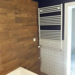 Wykończona łazienka z drewnopodobnymi panelami ściennymi, białymi płytkami, grzejnikiem drabinkowym i fragmentem wanny.