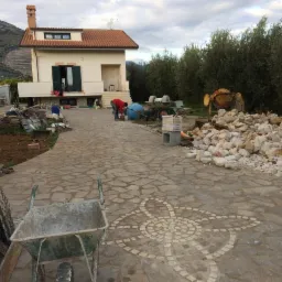Podjazd z kamienną nawierzchnią w trakcie budowy, z wzorem kwiatowym z kostki, w tle dom jednorodzinny, betoniarka i sterta kamieni.